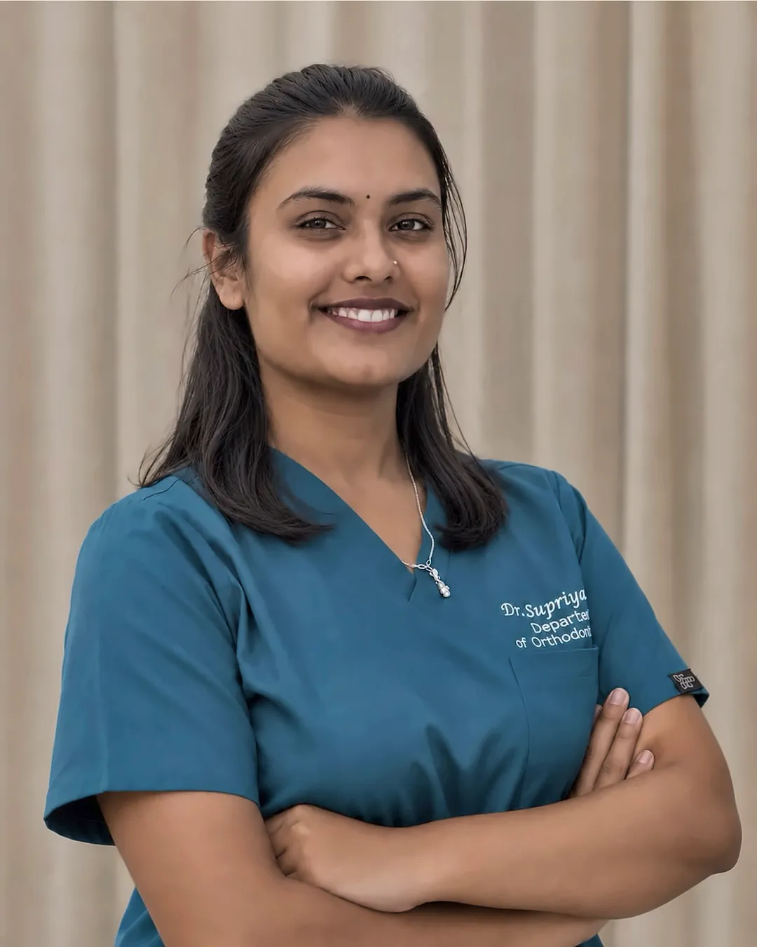 Dr. Supriya P, orthodontist, smiling with arms folded, in teal scrubs embroidered with her name